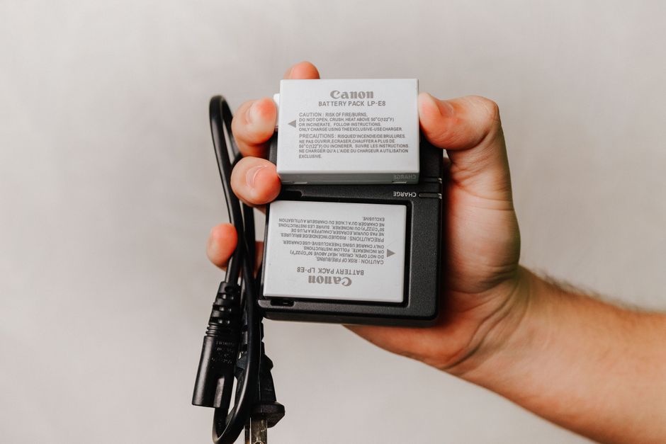A hand holding a Canon camera battery charger with two batteries, one labeled 'Canon Battery Pack LP-E8' and the other 'Canon Battery Pack LP-E8,' along with black charging cables. The background is a