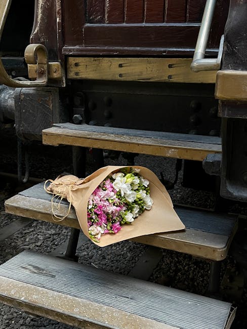 A vibrant bouquet of fresh flowers resting on a small wooden outdoor train station step, wrapped in brown kraft paper with a natural twine bow. The arrangement includes pink and white blooms, likely c