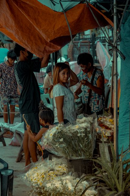 A scene at Dalston market under a large orange canopy features a flower stall with an assortment of fresh flowers, including white daisies and other colorful blooms, arranged in clear glass vases and 