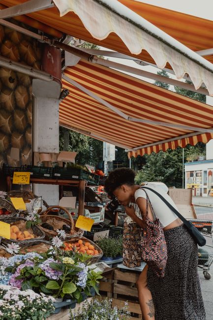 A woman with short dark hair, wearing glasses and a white top, is leaning forward to closely examine a floral display at Dalston market. She holds a patterned bag over one shoulder. The display featur