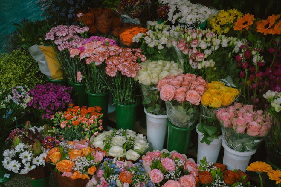 A vibrant display of fresh flowers in various arrangements and colours, including pink, white, yellow, orange, purple, and red blooms. The flowers are organized in green and white buckets, with some w
